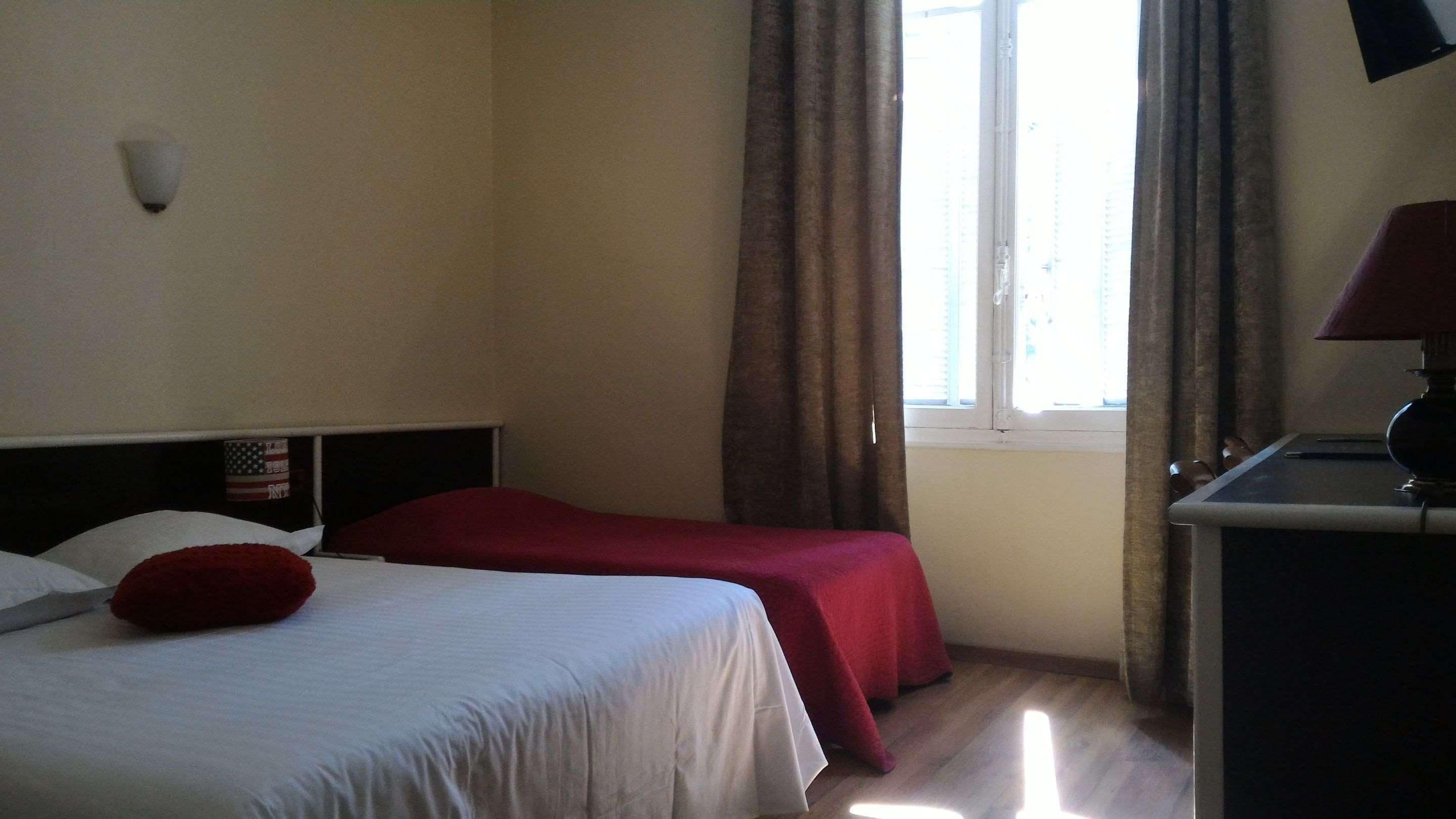 A hotel room with a bed, a desk, a lamp, a window with curtains, and a wall-mounted light.