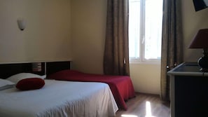 Una habitación de hotel con cama, escritorio, lámpara, ventana con cortinas y luz empotrada en la pared.