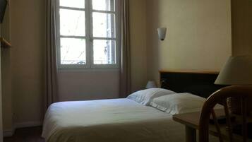 A hotel room with a bed, a chair, a window with blinds, and a wall-mounted lamp.