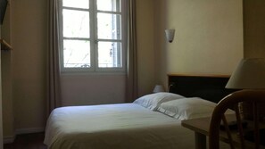 Une chambre d’hôtel avec un lit, une chaise, une fenêtre avec des stores et une lampe fixée au mur.