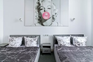 Quarto com duas camas, roupa de cama com padrão marmorizado, cabeceira cinza, mesa de cabeceira pequena e um quadro na parede.