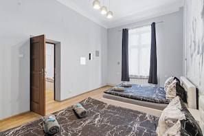A modern bedroom with a bed, a small seating area, a door leading to another room, and a window with curtains. - Queens Apartments (Kraków)