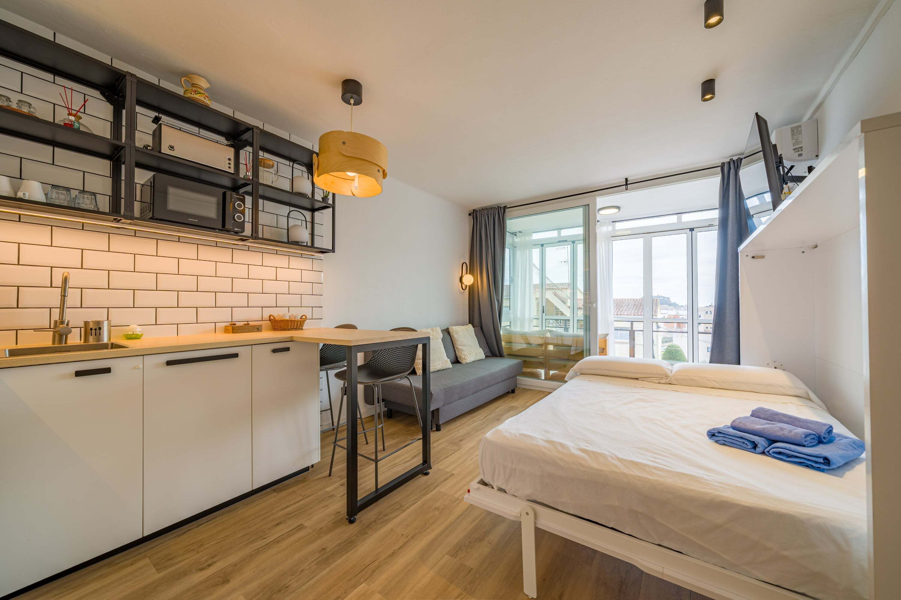 Une chambre d’hôtel compacte comprenant un coin cuisine, un lit avec des serviettes, un canapé et une grande fenêtre.