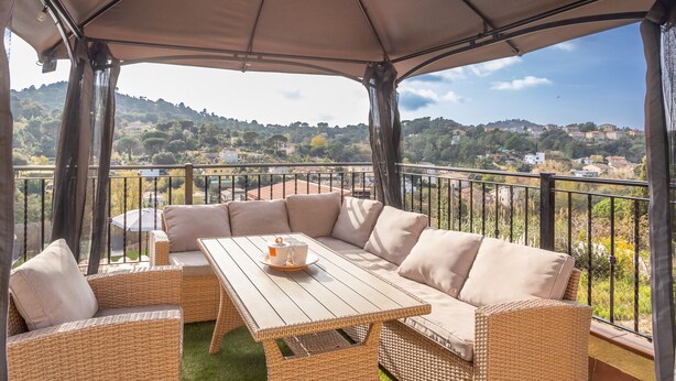 A covered outdoor seating area with wicker furniture and a table, overlooking a scenic hillside view. - Villa Luz Lloret with Pool (Lloret De Mar)