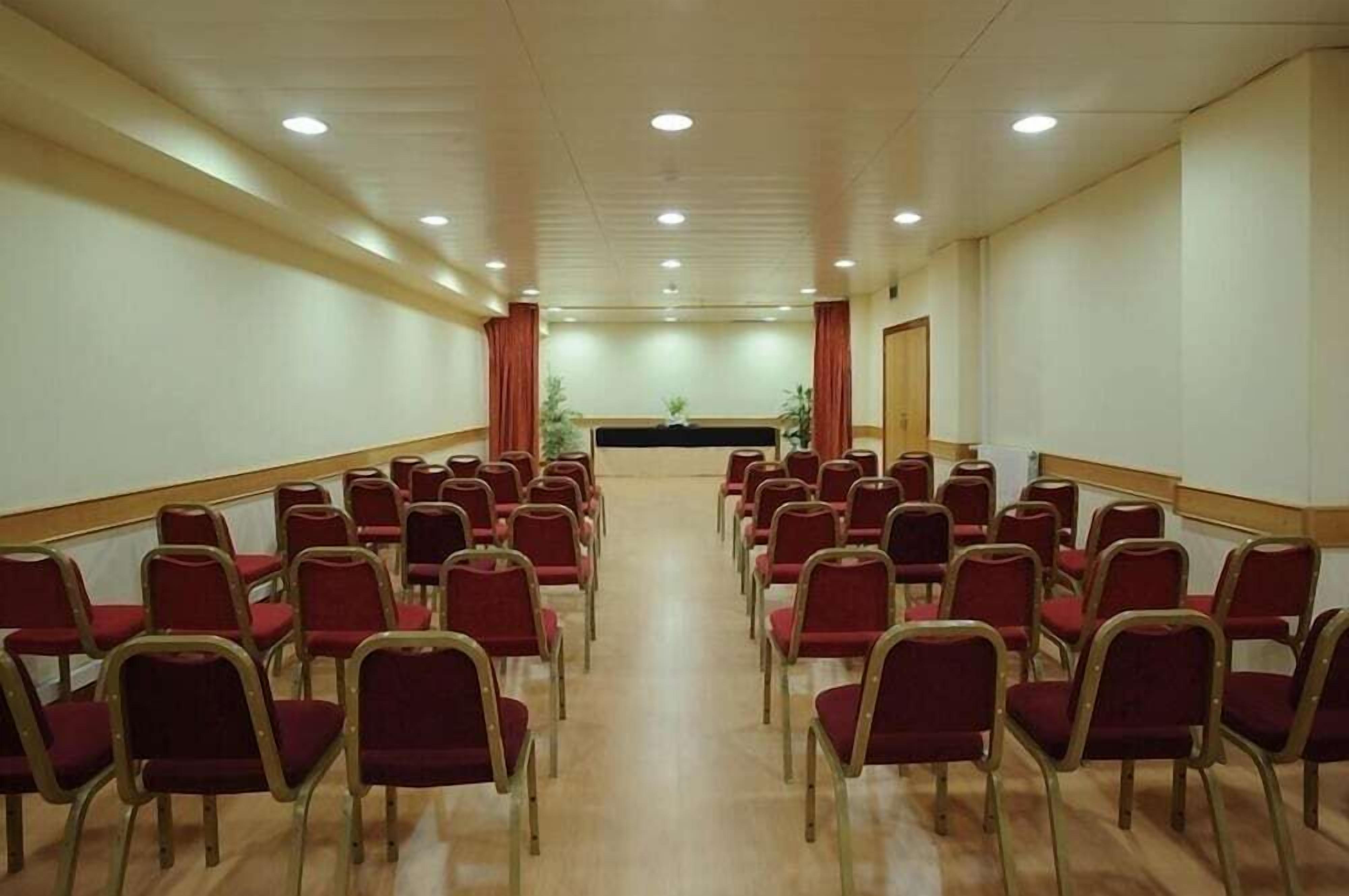 Une salle de conférence avec des rangées de chaises rouges, un pupitre à l’avant et une plante au centre.