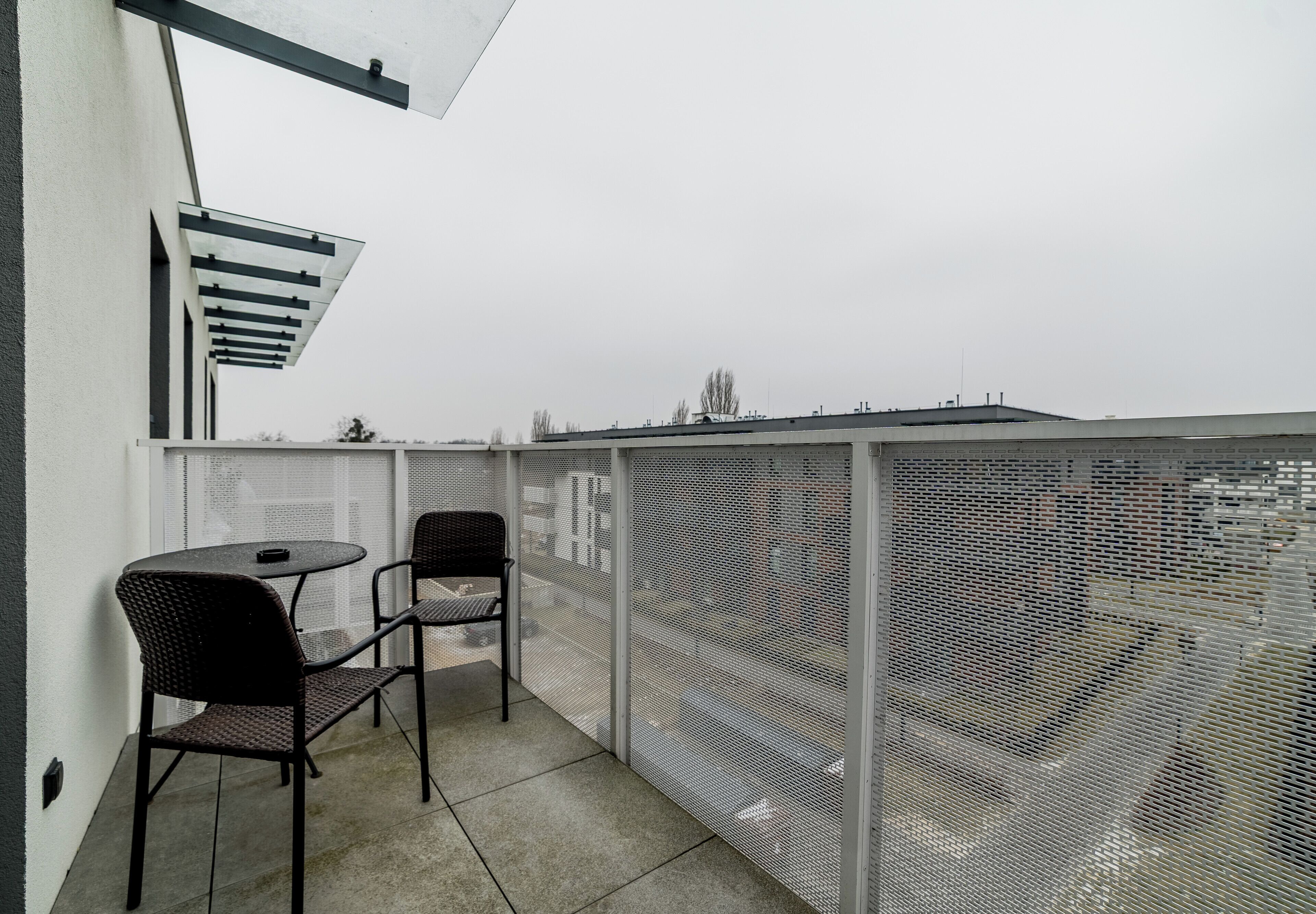 Uma varanda com duas cadeiras, uma mesa, grade de metal e vista para a cidade.