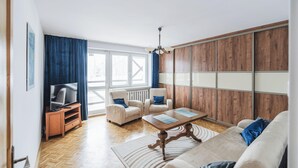 Een woonkamer met een bank, fauteuils, een salontafel, een televisie op een dressoir en een groot raam met gordijnen.