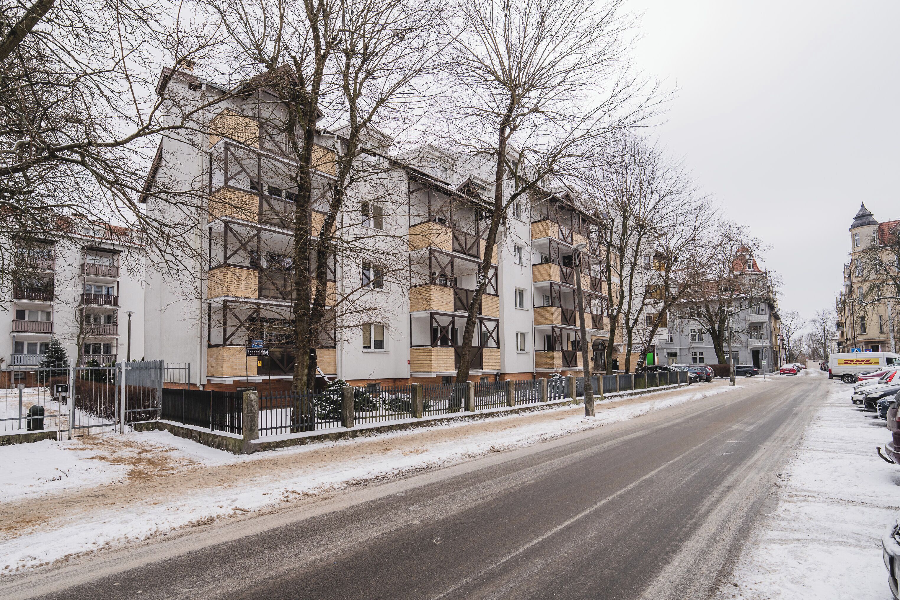 A multi-story building with balconies, a snow-covered street, and parked cars.