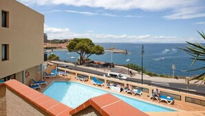 Zona de piscina con sillas reclinables azules y vista al mar y un fuerte.