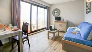 Habitación de hotel con un amplio ventanal corredero al balcón, un escritorio con una computadora portátil, una mesita con una lámpara, un sofá y un reloj de pared.