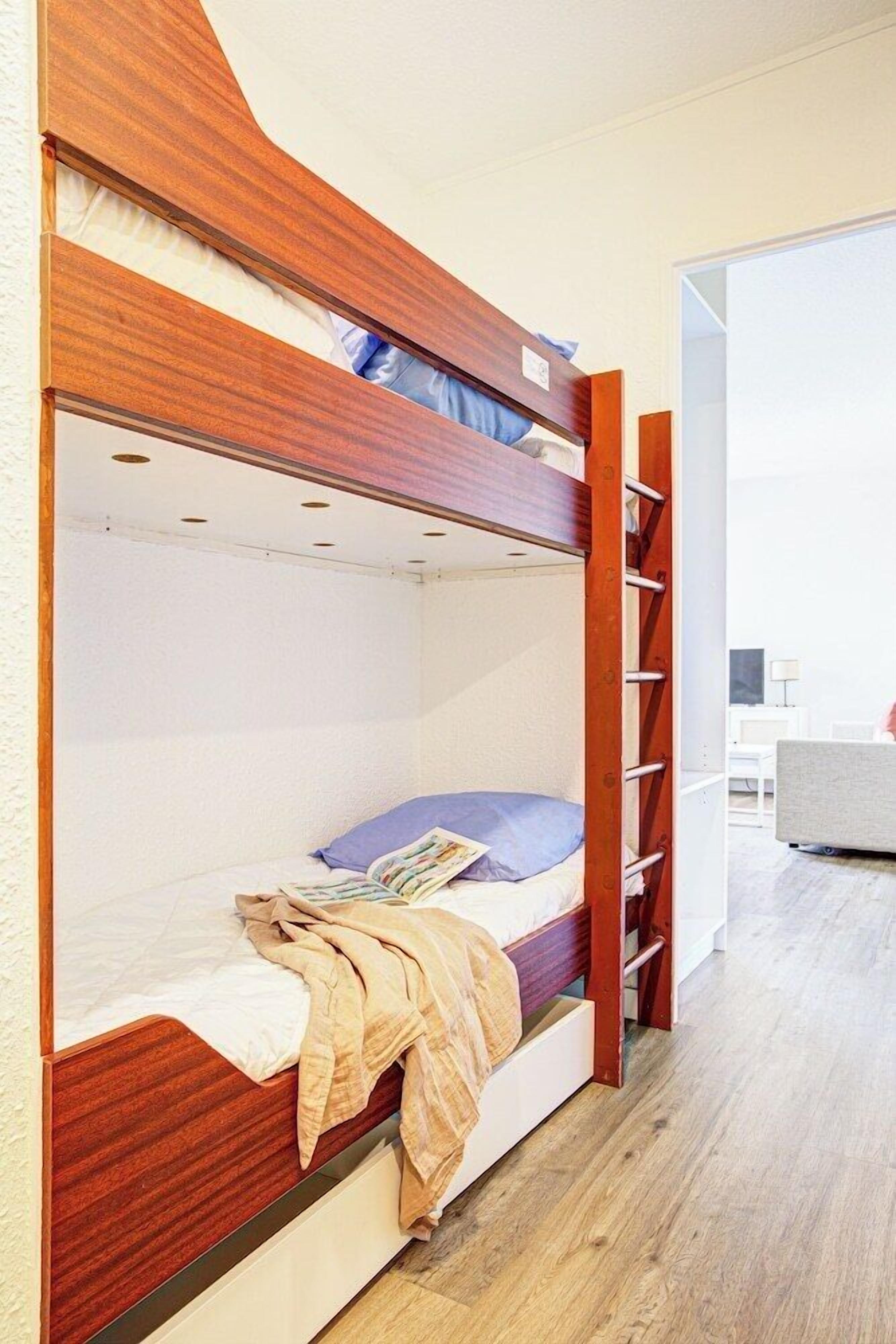 A bunk bed with a wooden frame, a white mattress, and a blue pillow. There is a wooden ladder attached to the side.