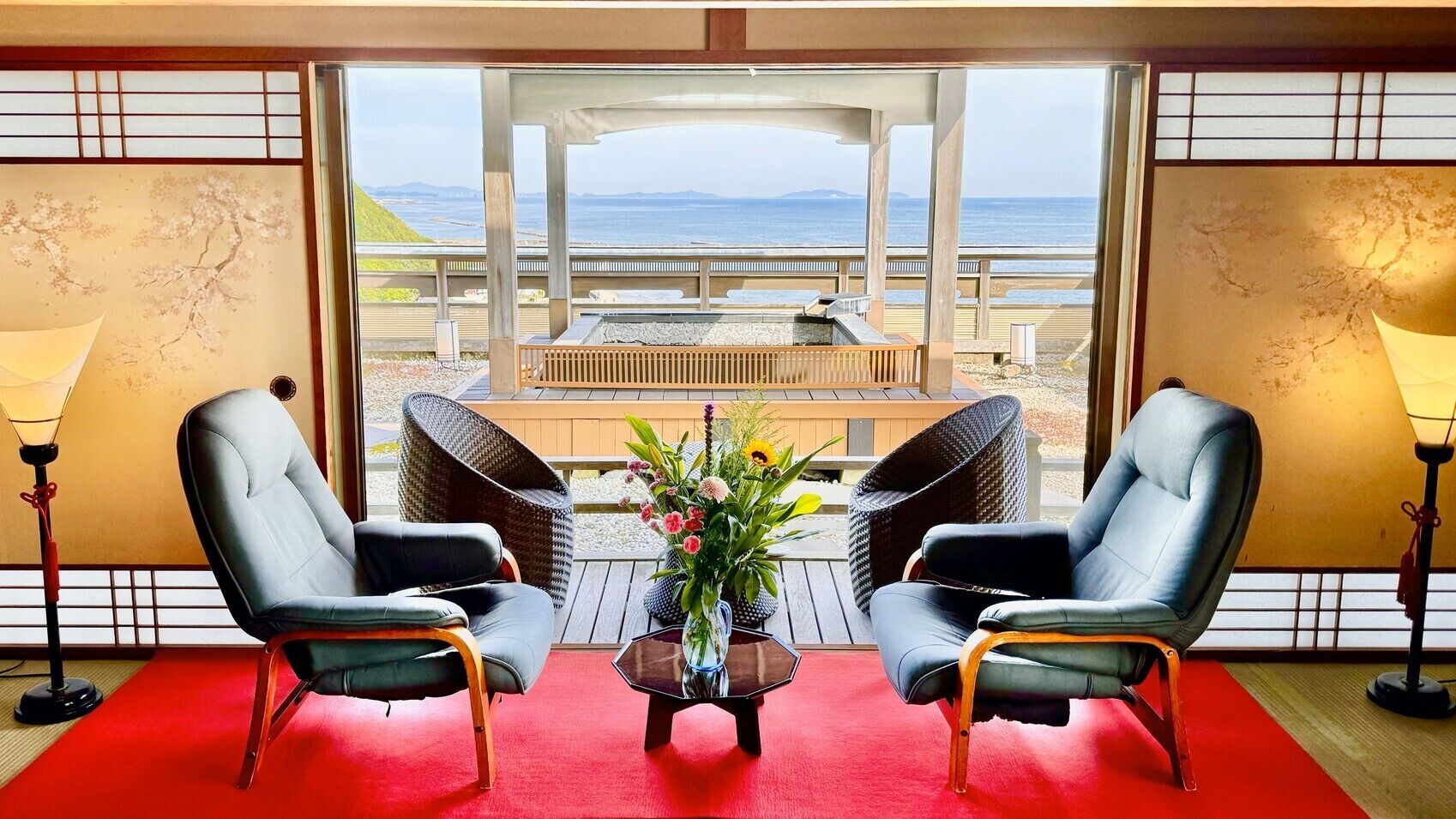 A room with two leather chairs, a small table with a vase of flowers, and a view of the beach and ocean through large windows.
