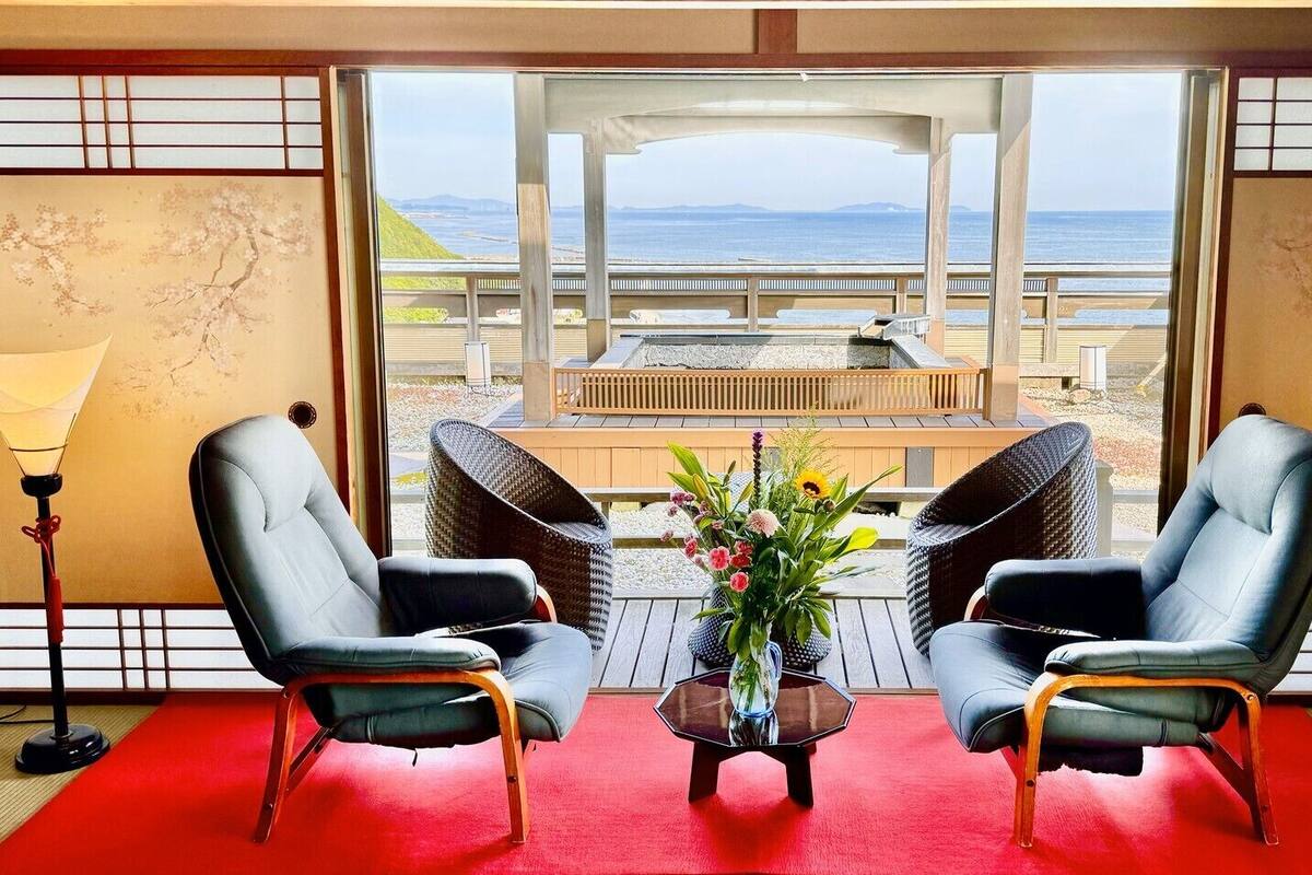 A room with two leather chairs, a small table with a vase of flowers, and a view of the beach and ocean through large windows.