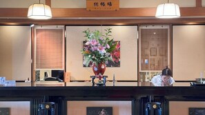 A reception desk with a vase of flowers, a wooden sign above, and a person in the background. - Shukukaifu (Minamichita)