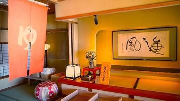 Une pièce de style japonais traditionnel, avec un sol recouvert de tatamis, un paravent en papier de riz (shoji), une œuvre de calligraphie, un vase de fleurs et une petite table avec une lampe.