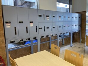 A row of grey lockers with numbered labels, a wooden table, and chairs in a room with tiled flooring. - Toyoko Inn Himeji Eki Shinkansen Minami Guchi (Himeji)