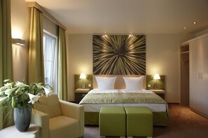 A hotel room with a large bed, two bedside lamps, a green upholstered bench, a potted plant, and a decorative wall piece. - The Originals Kur Und Wellnesshaus Spreebalance (Burg (Spreewald))