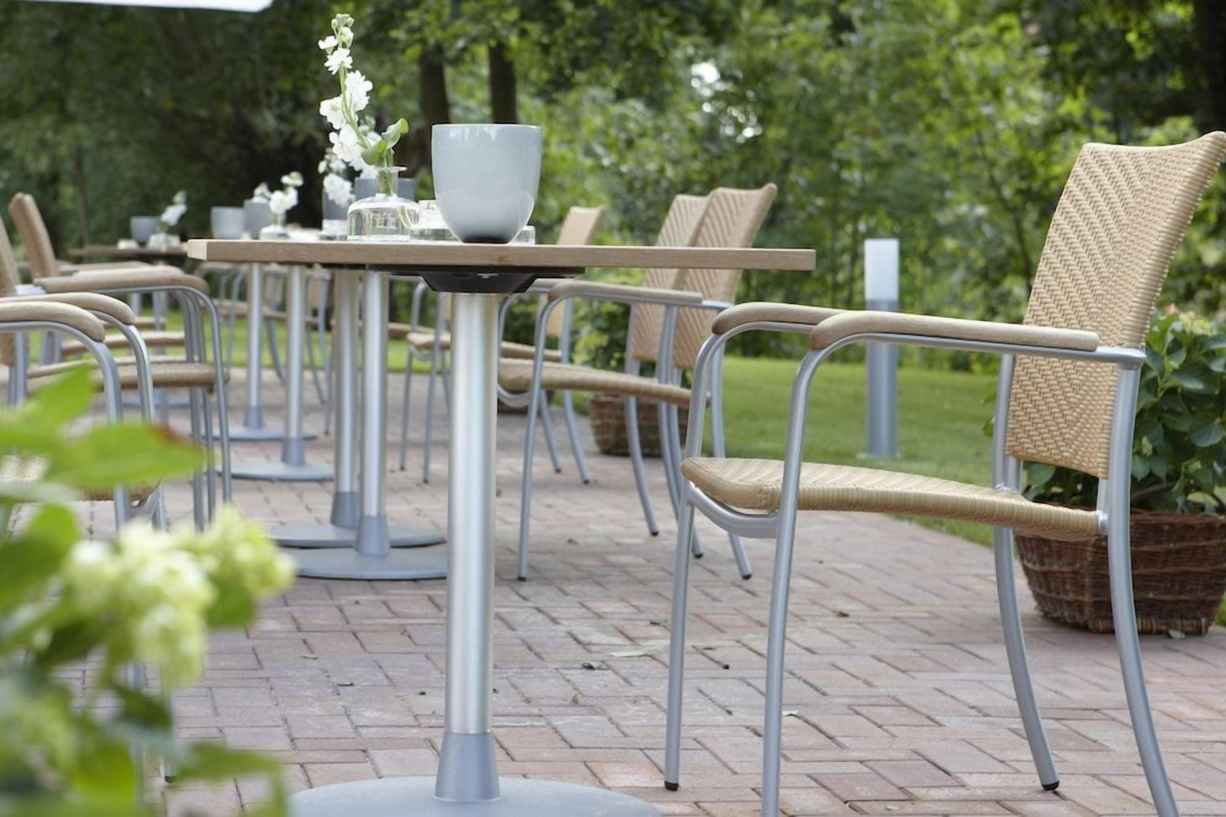 Terrasse mit Sitzgelegenheiten, Metalltische und Korbstühle, umgeben von viel Grün.
