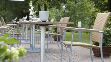 Terrasse mit Sitzgelegenheiten, Metalltische und Korbstühle, umgeben von viel Grün.