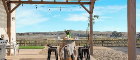 A covered outdoor seating area with a table set for two, surrounded by chairs, under a wooden pergola with string lights.