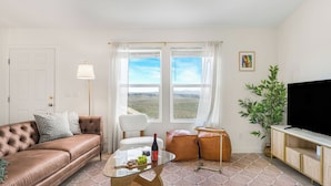 A living room with a brown leather sofa, a glass coffee table with a bottle and grapes, a white chair, a large window with a view of the countryside, and a TV on a wooden stand. - Bombay Chardonnay by Avantstay Covered Pavilion + BBQ Quiet Paso Hillside + Vineyards View (Paso Robles)