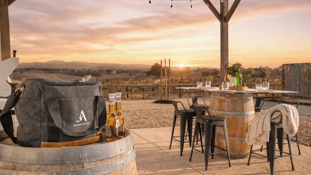 A table set outdoors with a view of the countryside, a barrel, and a bag with 'AVANTSTAY' on it. - Bombay Chardonnay by Avantstay Covered Pavilion + BBQ Quiet Paso Hillside + Vineyards View (Paso Robles)