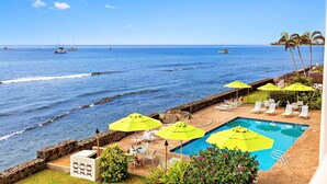 A pool area with yellow umbrellas, a stone wall, and a view of the ocean with boats. - New Listing! Ocean Front Gem in Lahaina (Lahaina)