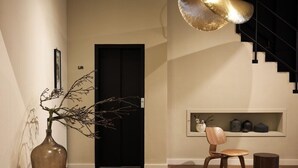 A modern interior with a wooden floor, a black door labeled "Lift", a wooden chair, a small round table, and a wall-mounted shelf with decorative items. - St. Martenslane Hotel Maastricht (Maastricht)