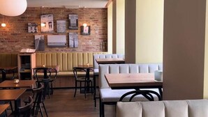 A restaurant interior with wooden tables and chairs, a brick wall with framed pictures, and a large window on the right. - St. Martenslane Hotel Maastricht (Maastricht)