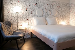 A modern bedroom with a geometric patterned wall, a bed with white bedding, a tufted armchair, and two bedside lamps. - Mabi Hotel Centrum (Maastricht)