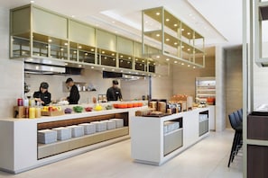 A modern kitchen with chefs preparing food, a buffet counter with various food items, and a seating area with a bar. - Courtyard By Marriott Osaka Honmachi (Osaka)