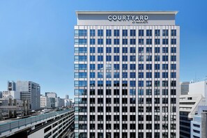A Courtyard by Marriott hotel with a modern glass facade, surrounded by urban buildings and a covered parking structure. - Courtyard By Marriott Osaka Honmachi (Osaka)