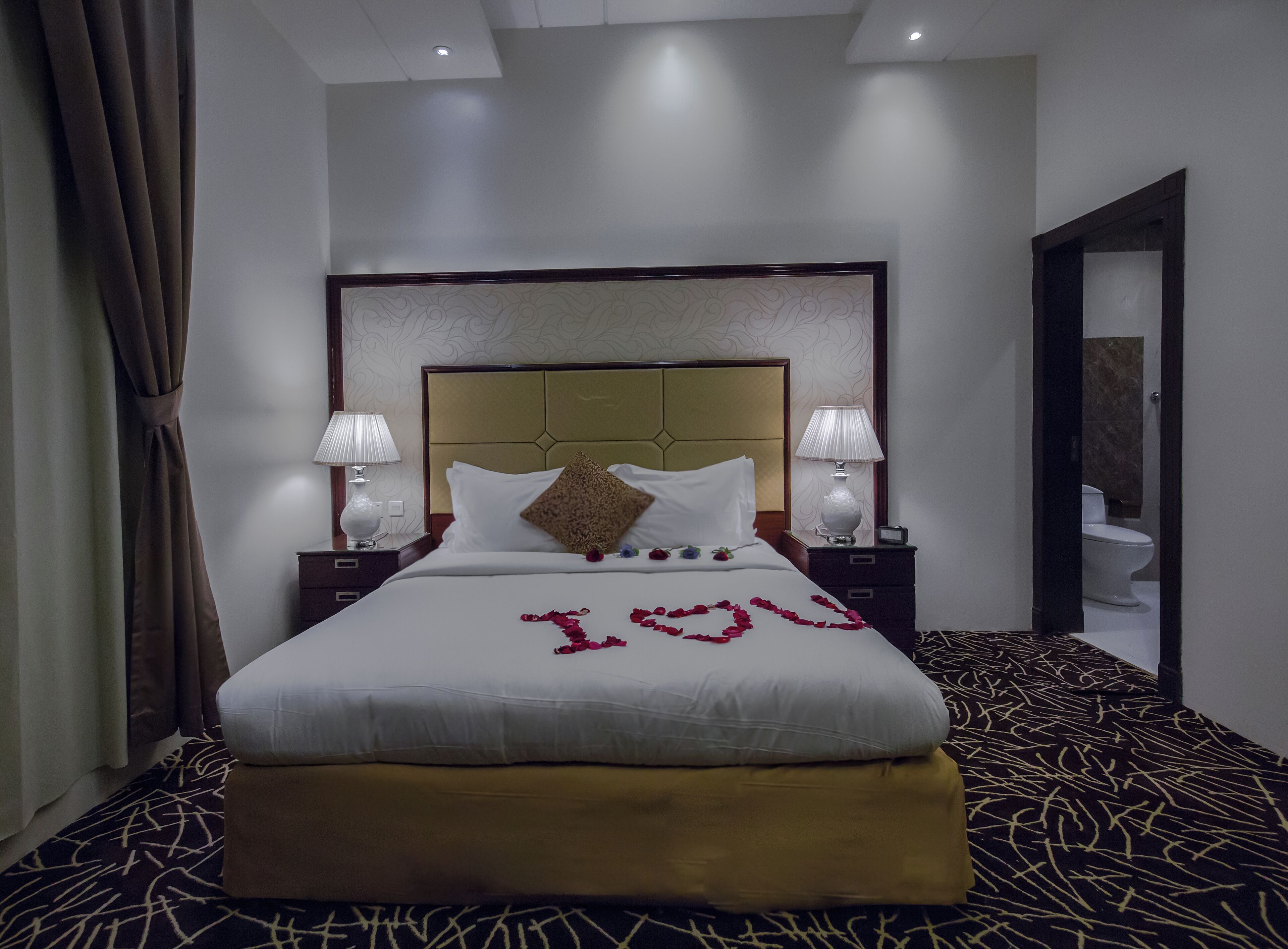 A hotel room with a large bed, two bedside lamps, a headboard, and a bathroom visible through an open door.