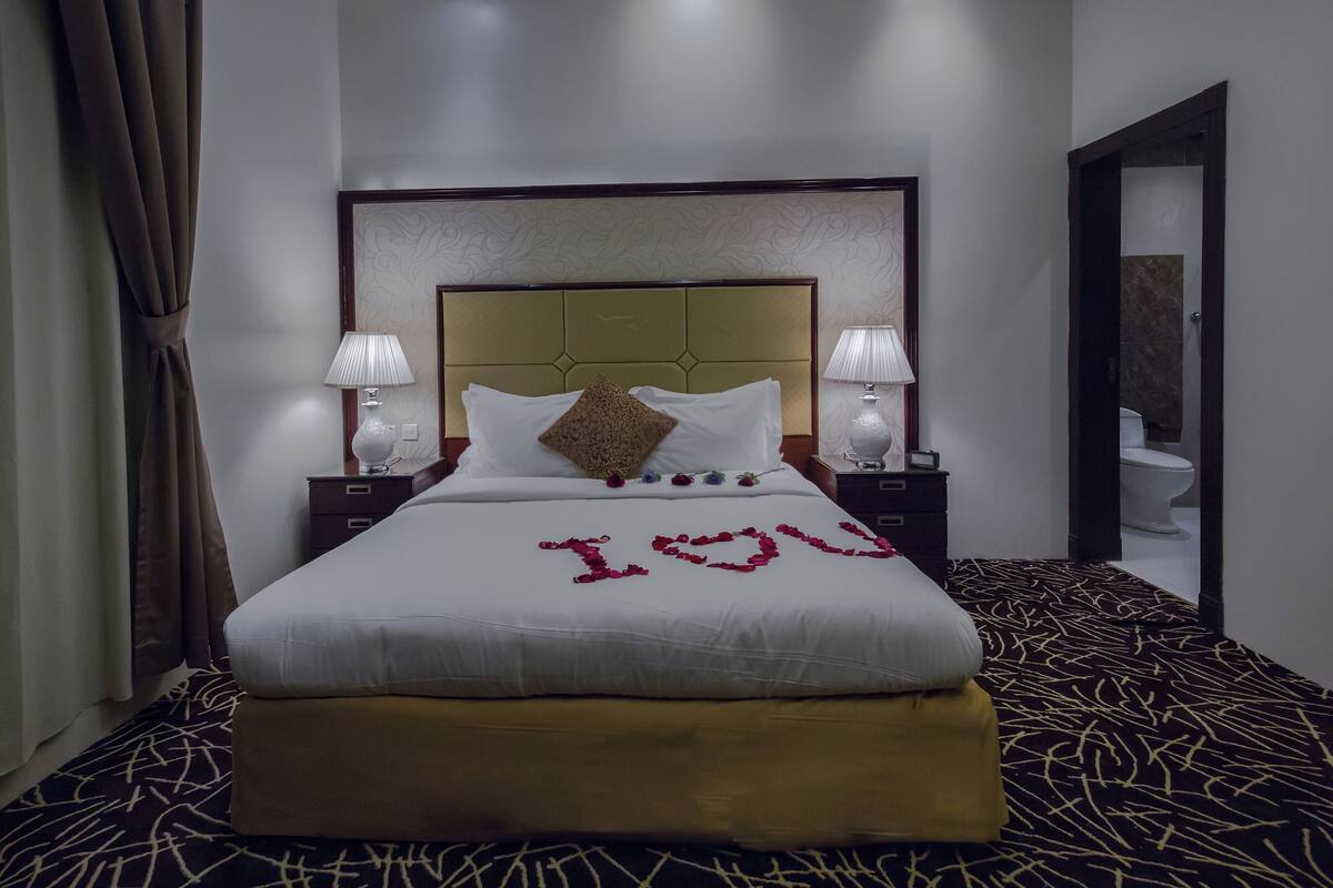 Ein Hotelzimmer mit einem großen Bett, zwei Nachttischlampen, einer Kopfteil und einem durch eine offene Tür einsehbaren Badezimmer.