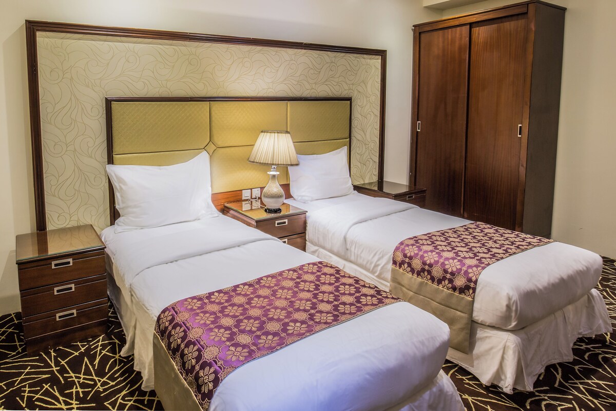Habitación de hotel con dos camas, una mesita de noche con lámpara, un armario y una alfombra estampada.