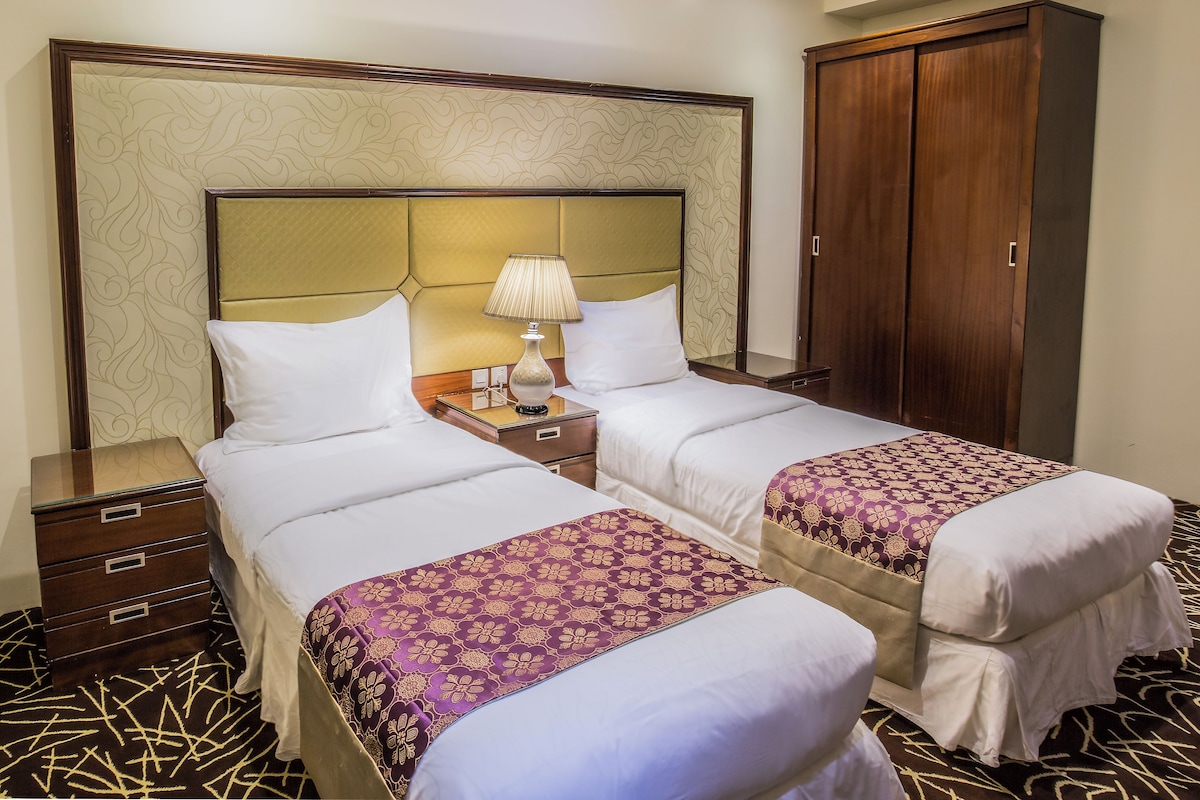 A hotel room with two beds, a nightstand with a lamp, a wardrobe, and a patterned carpet.