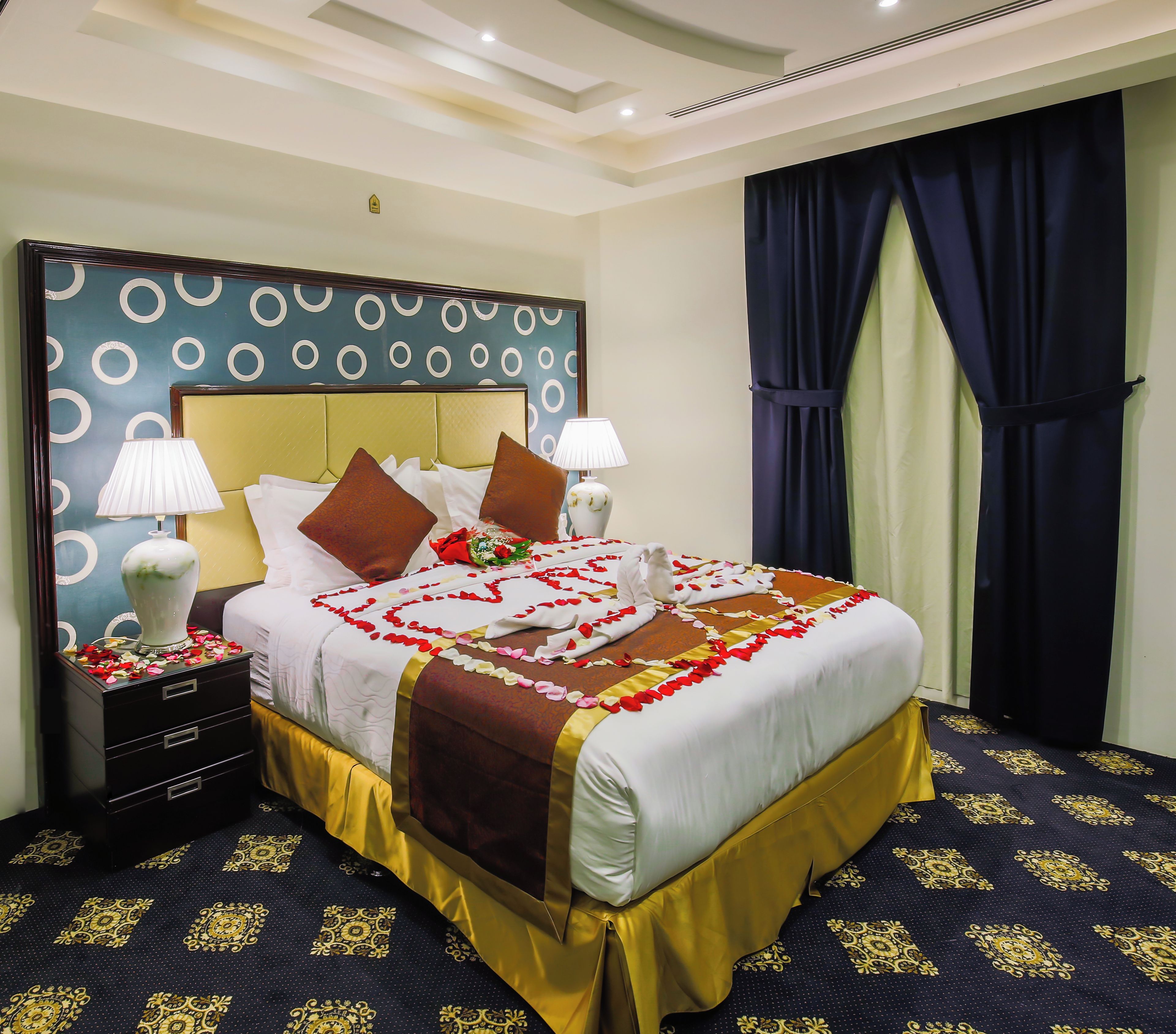 A hotel room with a large bed, two bedside tables, a nightstand, a headboard with a circular pattern, and a window with curtains.