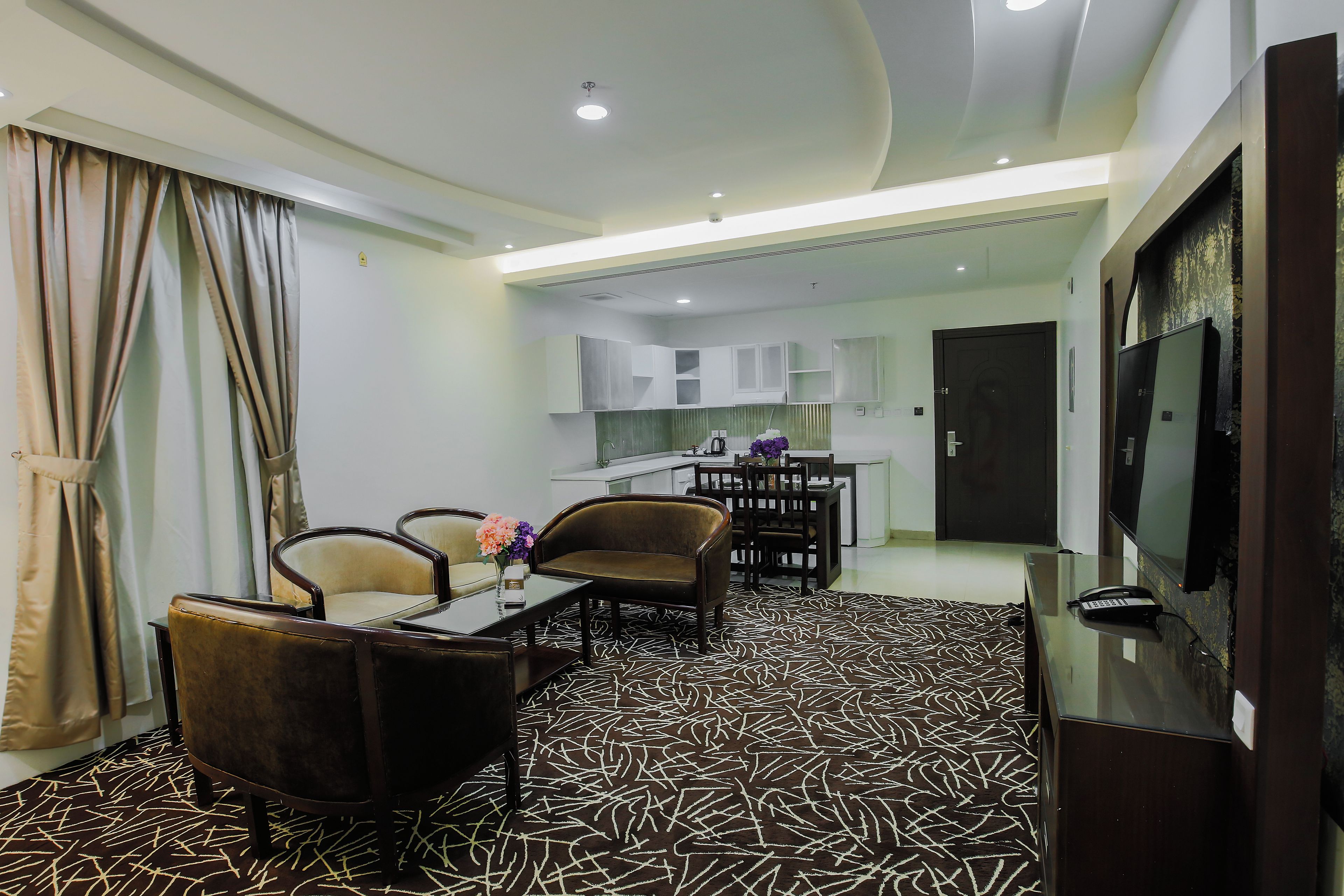 A modern living room with a sofa, chairs, a coffee table, and a TV. There is a kitchen area with cabinets and a dining table in the background.