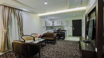 A modern living room with a sofa, chairs, a coffee table, and a TV. There is a kitchen area with cabinets and a dining table in the background.