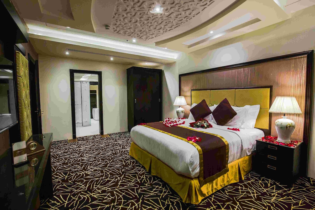 A hotel room with a large bed, two bedside tables with lamps, a TV, and a patterned carpet.