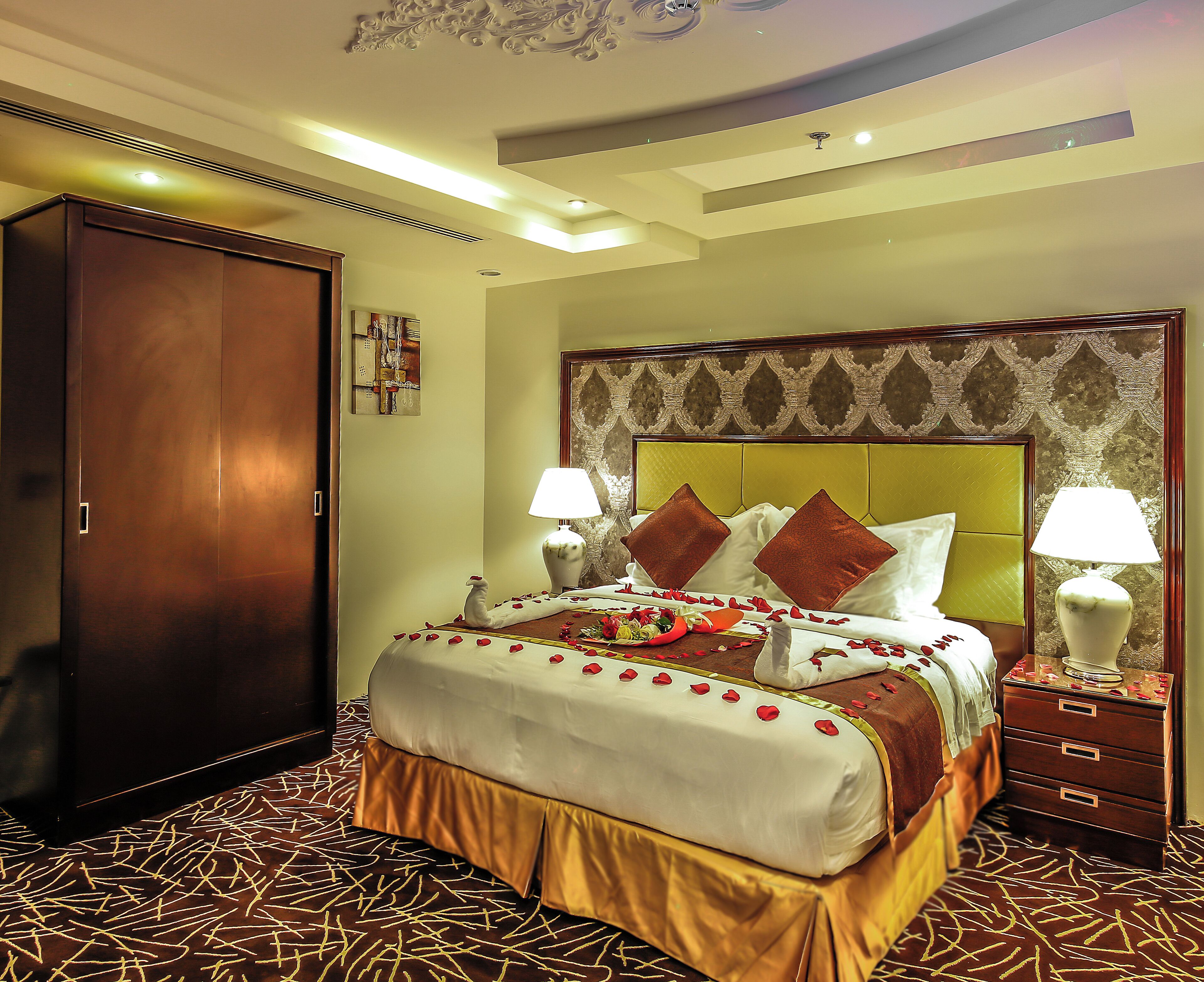 A hotel room with a large bed, two bedside tables with lamps, a wardrobe, and a decorative ceiling.