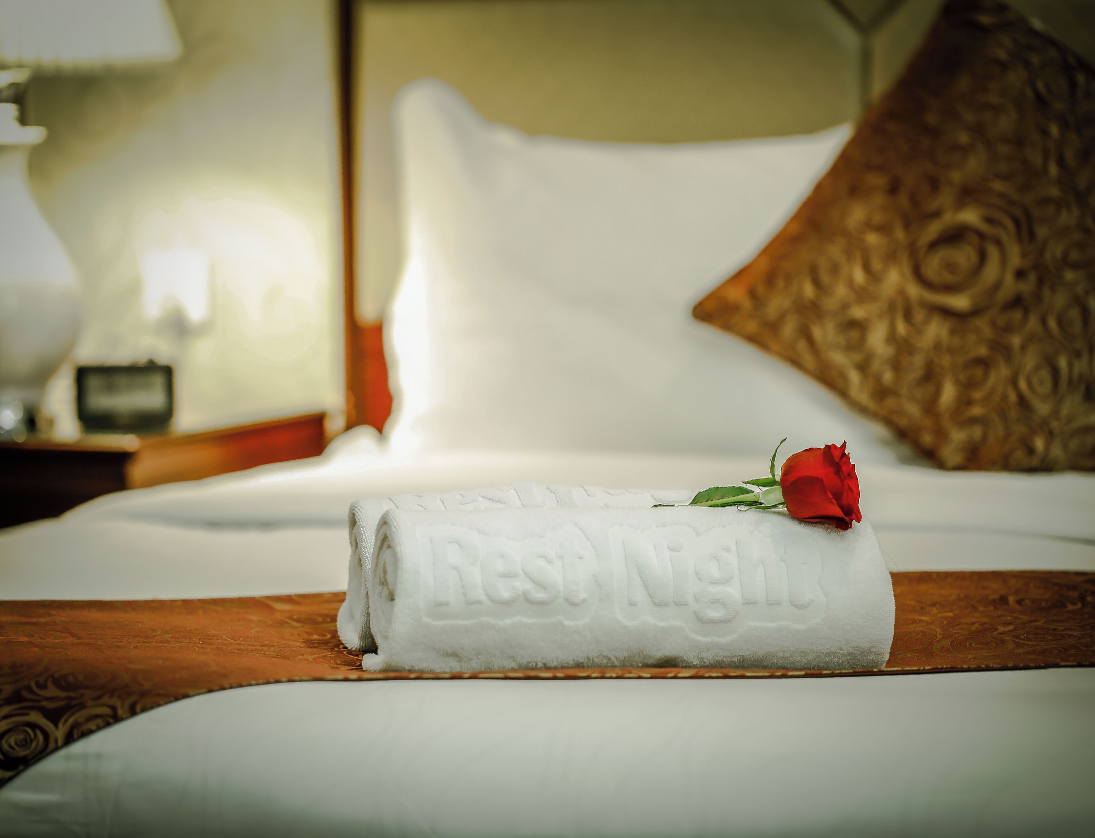 A neatly made bed with a folded towel and a rose, a wooden headboard, and a patterned pillow.