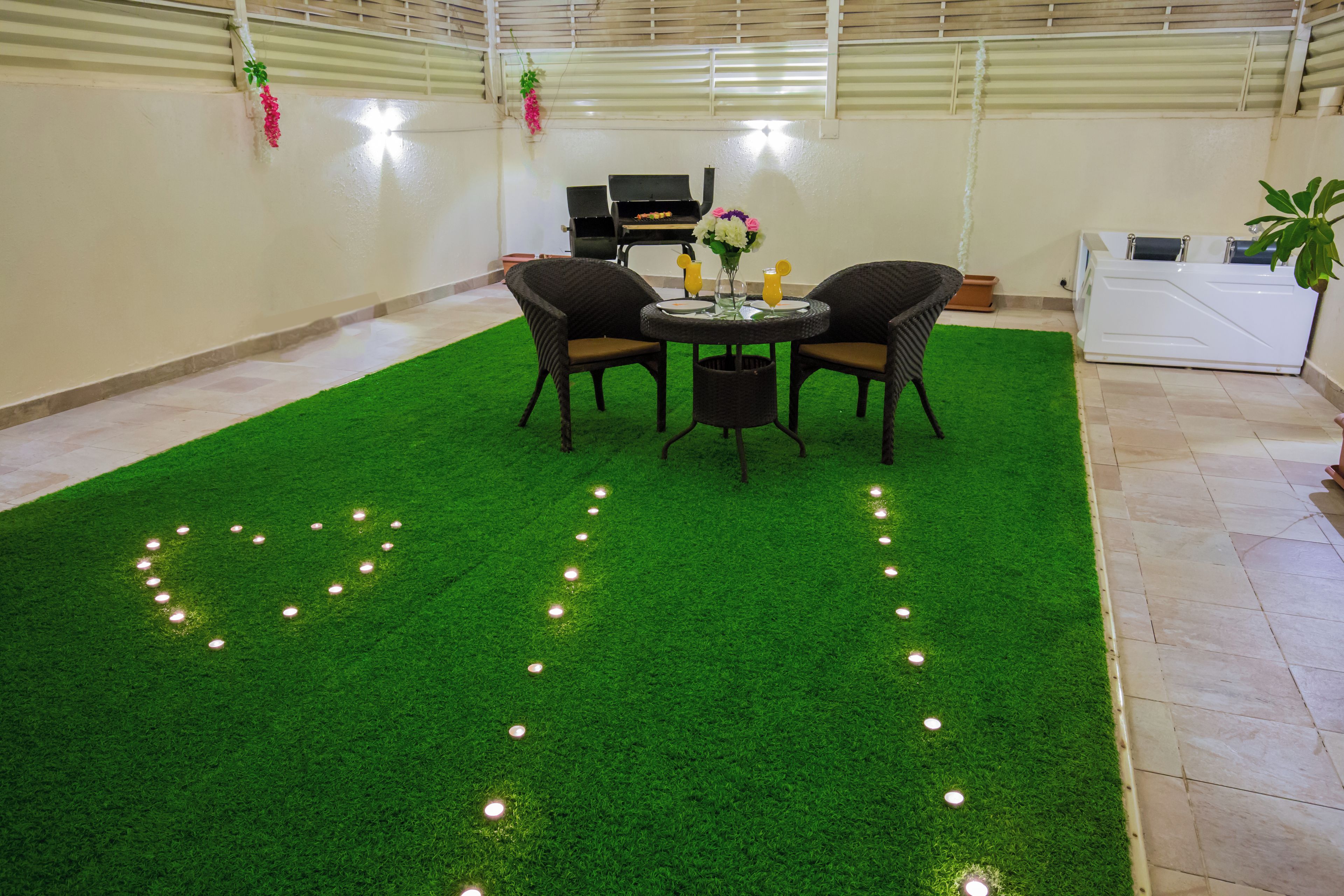 A room with a green artificial turf floor, a round table with chairs, a grill, and a white washing machine.