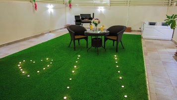A room with a green artificial turf floor, a round table with chairs, a grill, and a white washing machine.