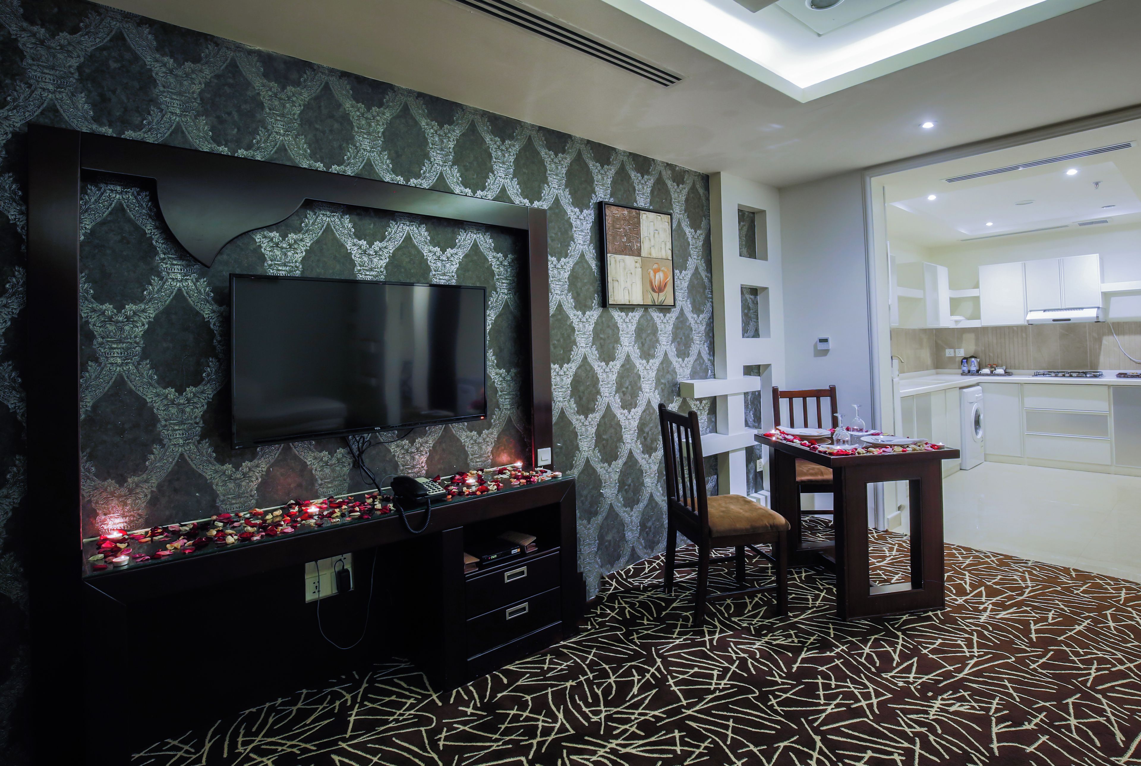 A modern living room with a flat-screen TV, a fireplace with candles, a dining table with chairs, and a patterned carpet.
