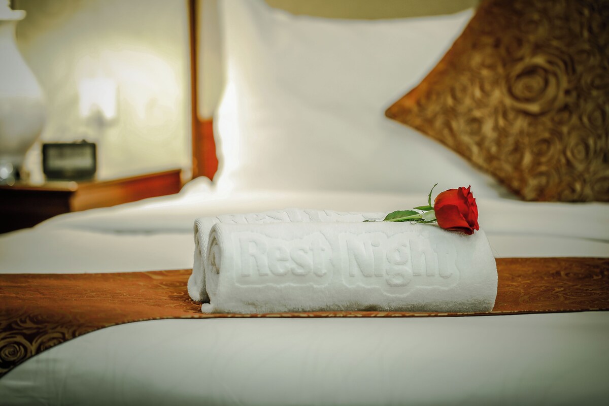 A neatly made bed with a folded towel and a rose, a wooden headboard, and a patterned pillow.
