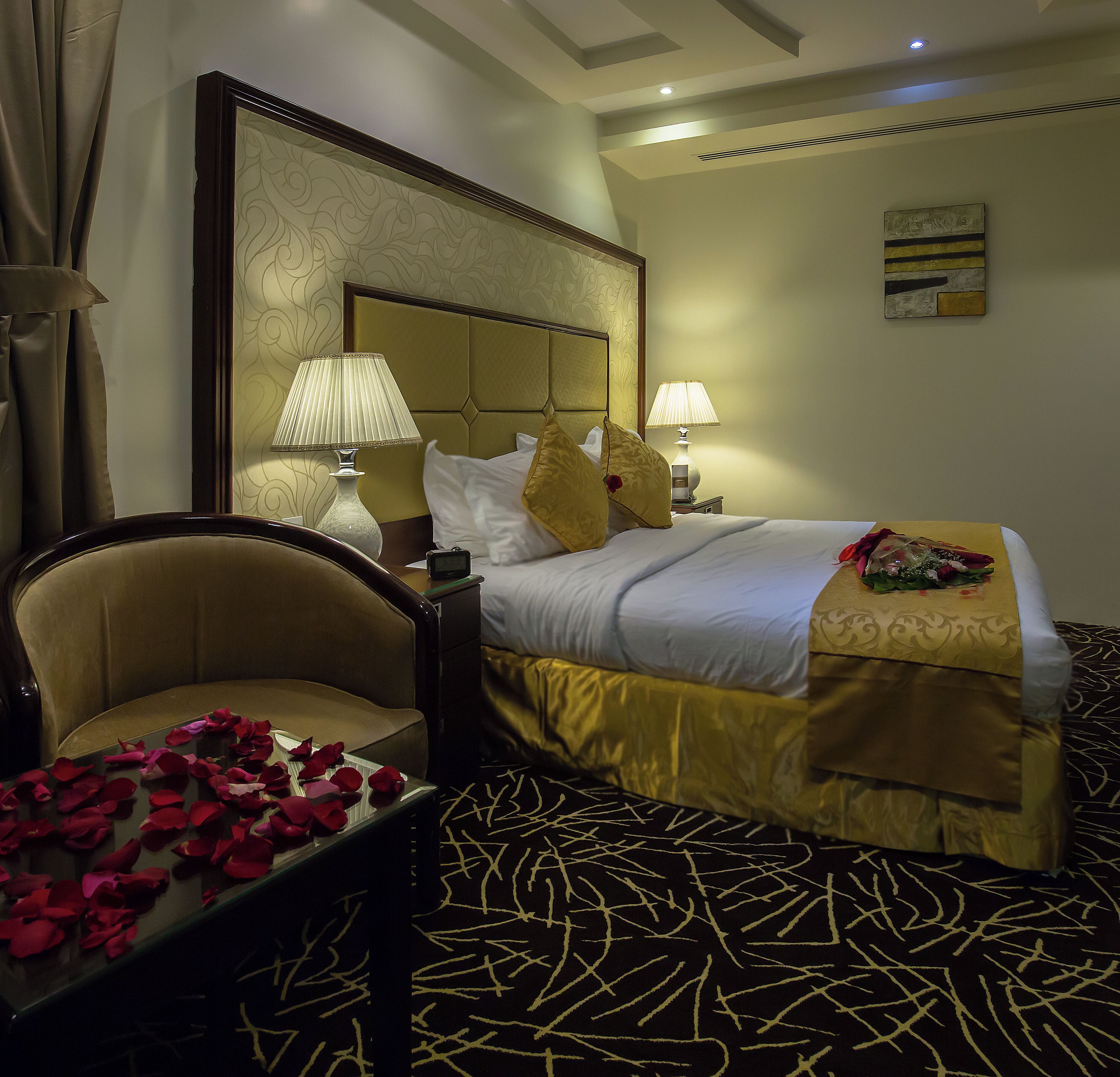 A hotel room with a bed, two bedside lamps, a chair, and a table with rose petals.