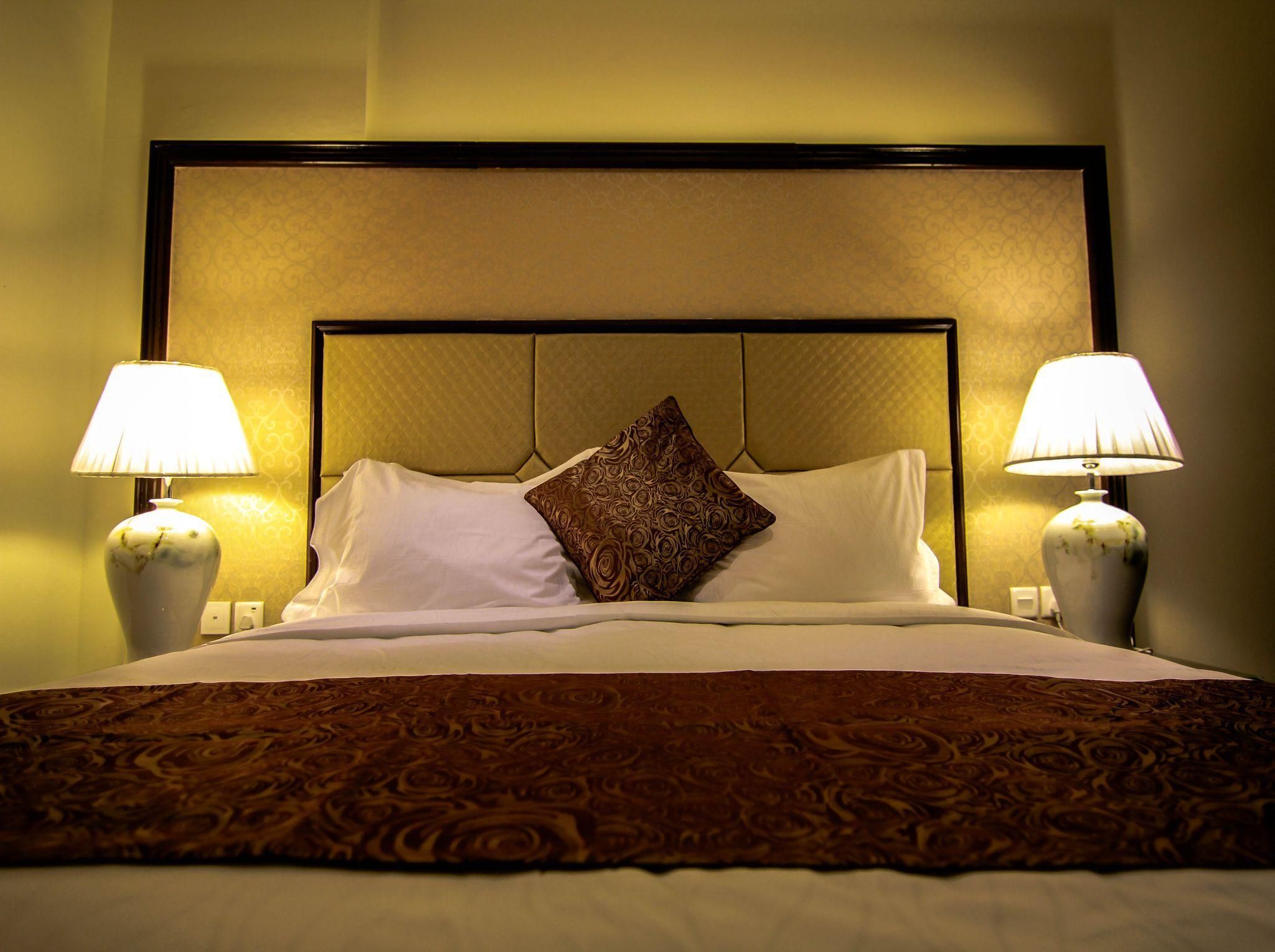 A hotel room with a bed, two bedside lamps, a patterned throw blanket, and a decorative pillow.