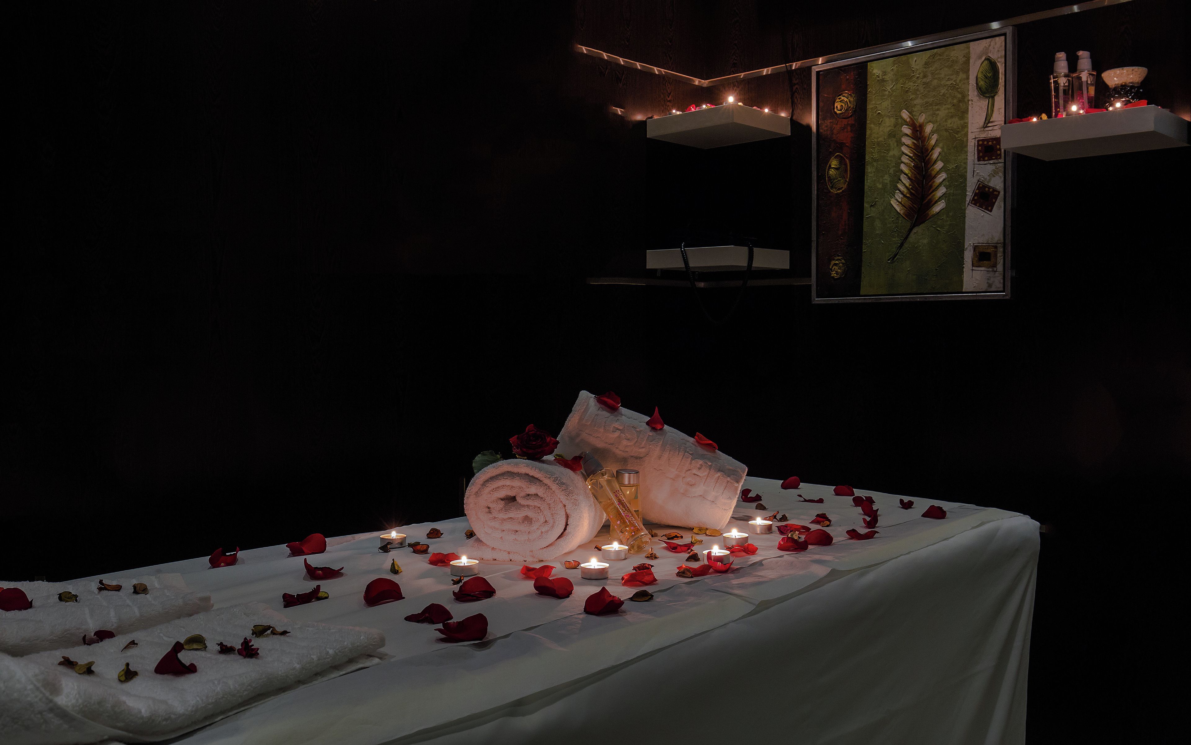 A dimly lit room with a bed covered in white sheets, surrounded by rose petals and lit candles.