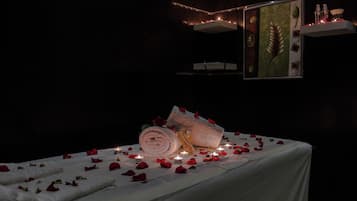 A dimly lit room with a bed covered in white sheets, surrounded by rose petals and lit candles.